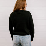 Back view of woman wearing a black sweater with a playing card graphic design featuring a red cowgirl and sequin hearts against a white background. 