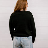 Back view of woman wearing a black sweater with a playing card graphic design featuring a red cowgirl and sequin hearts against a white background. 