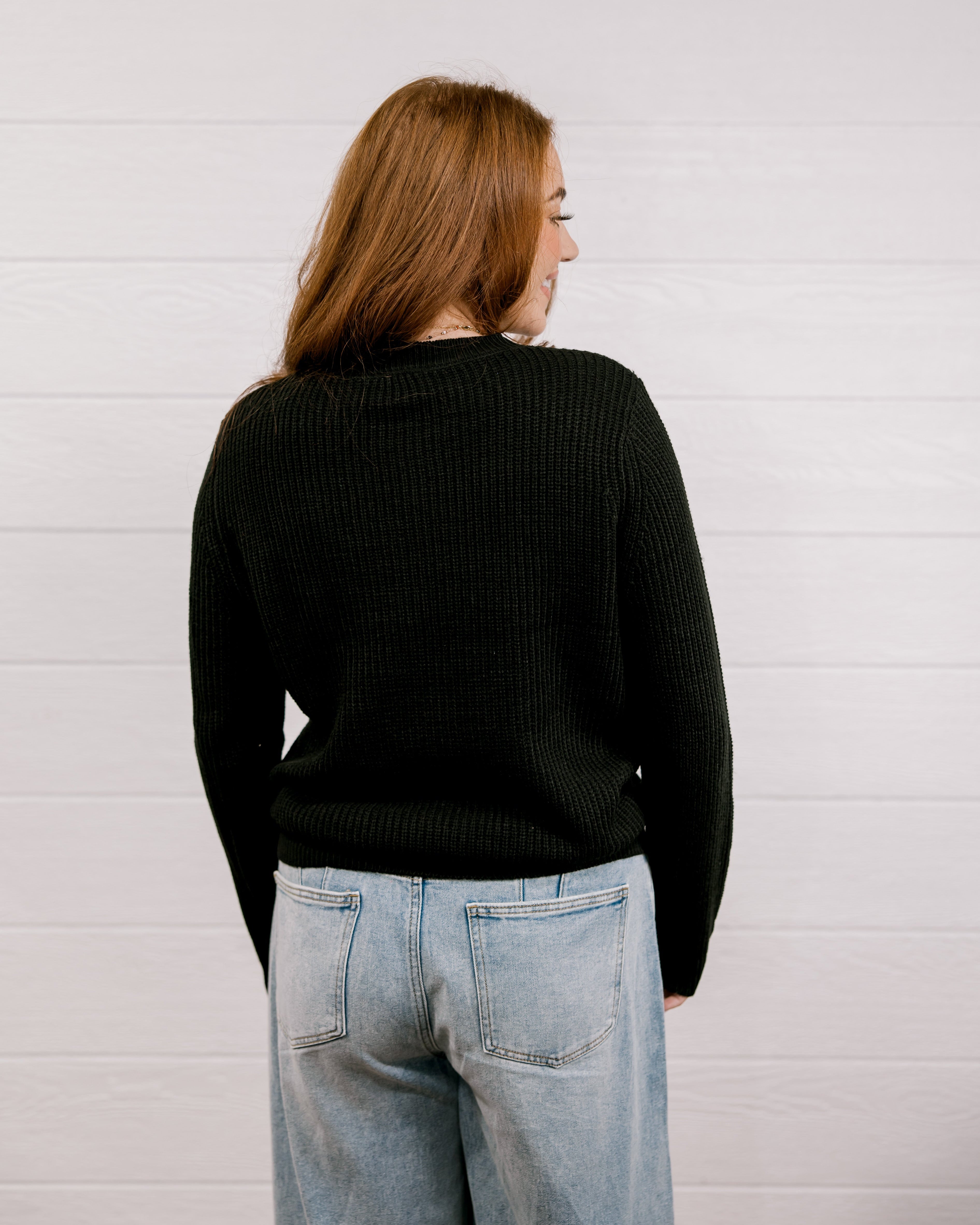 Back view of woman wearing a black sweater with a playing card graphic design featuring a red cowgirl and sequin hearts against a white background. 