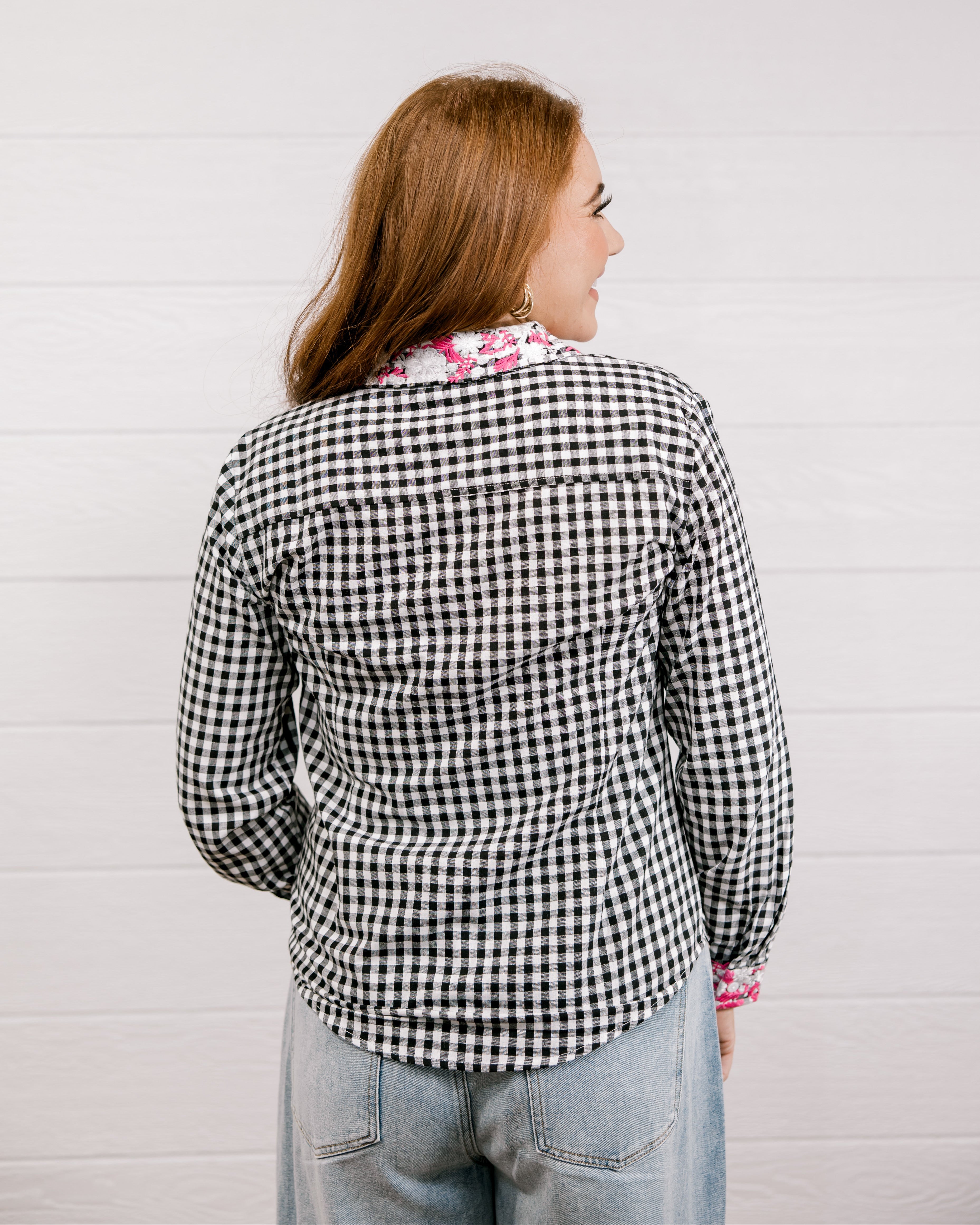 Back view of woman wearing a black and white checkered shirt with pink & white embroidery at collar and wrists against a white background.