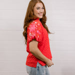 Side view of woman wearing a red textured top with embroidery on puff sleeves against a white background.