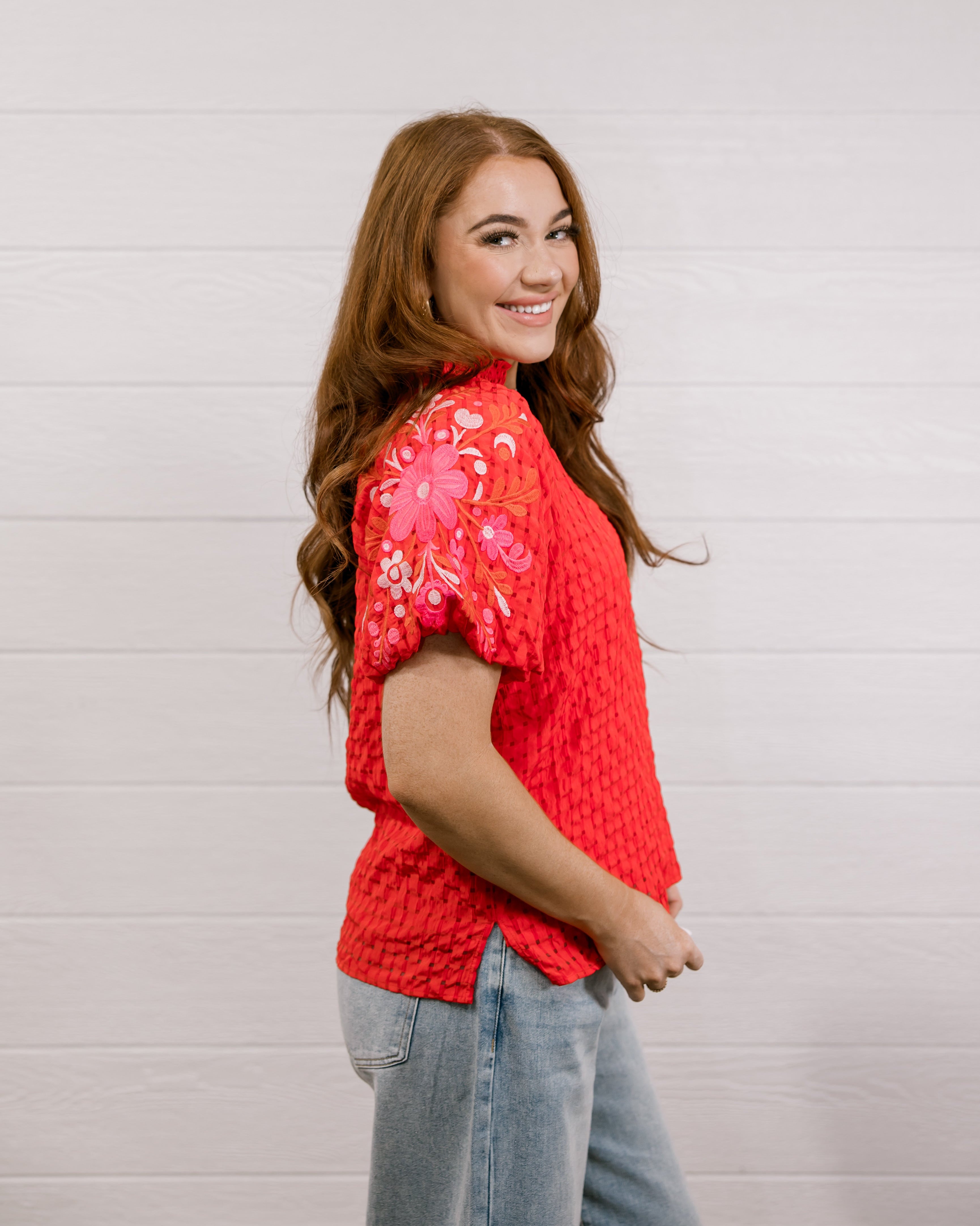 Side view of woman wearing a red textured top with embroidery on puff sleeves against a white background.