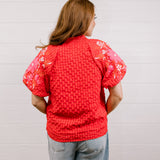 Back view of woman wearing a red textured top with embroidery on puff sleeves against a white background.