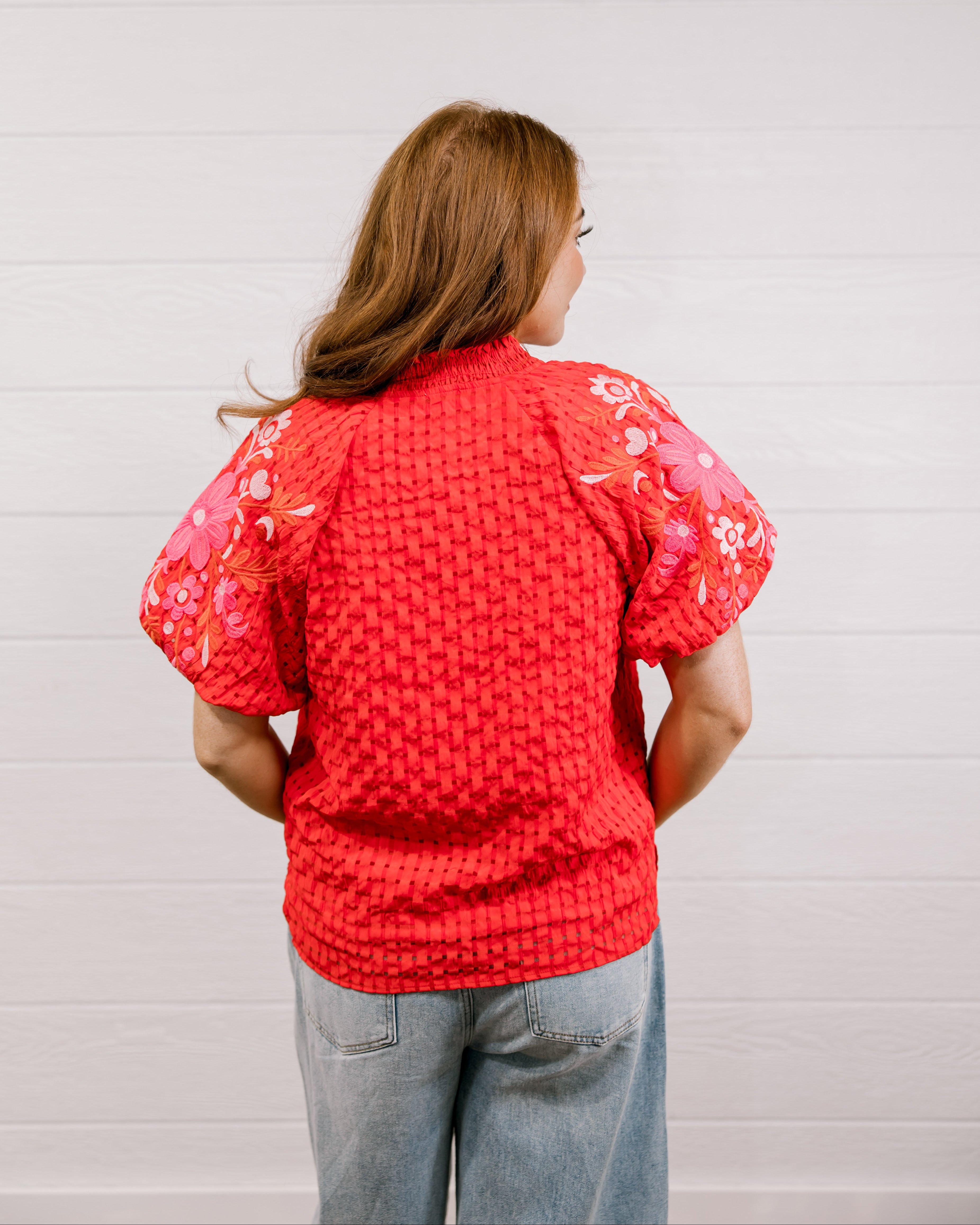 Back view of woman wearing a red textured top with embroidery on puff sleeves against a white background.