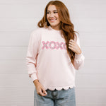 Woman wearing a pink sweatshirt with 'XOXO' embroidered design with scalloped hemline against a white background.