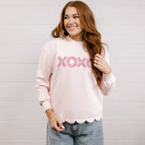 Woman wearing a pink sweatshirt with 'XOXO' embroidered design with scalloped hemline against a white background.