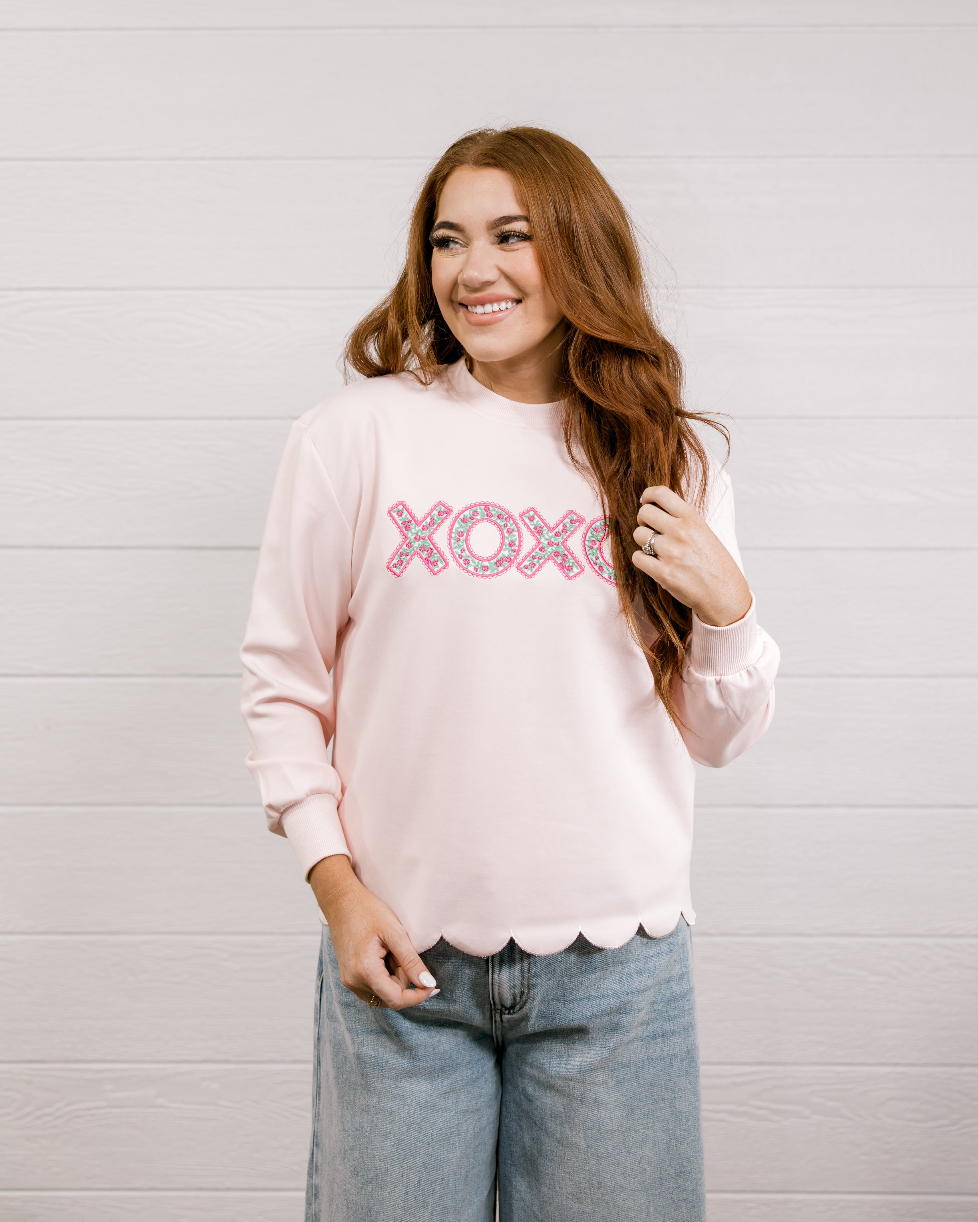 Woman wearing a pink sweatshirt with 'XOXO' embroidered design with scalloped hemline against a white background.