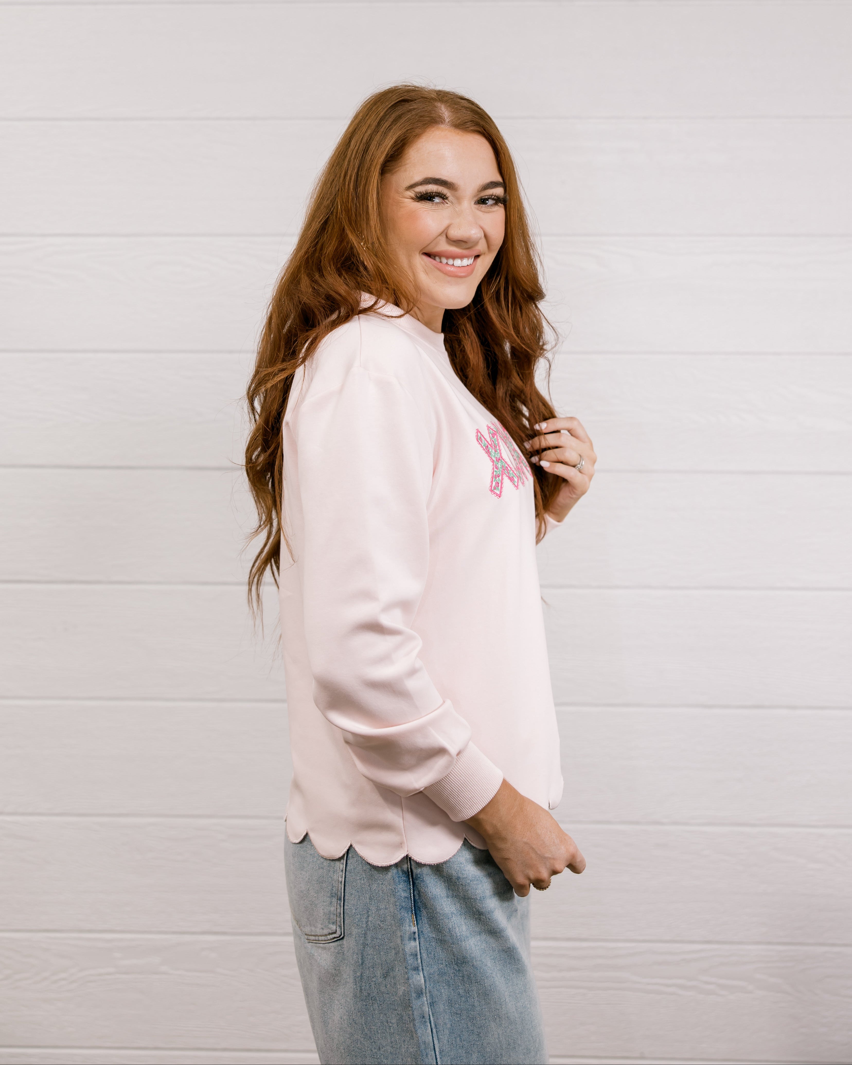 Side view of woman wearing a pink sweatshirt with 'XOXO' embroidered design with scalloped hemline against a white background.
