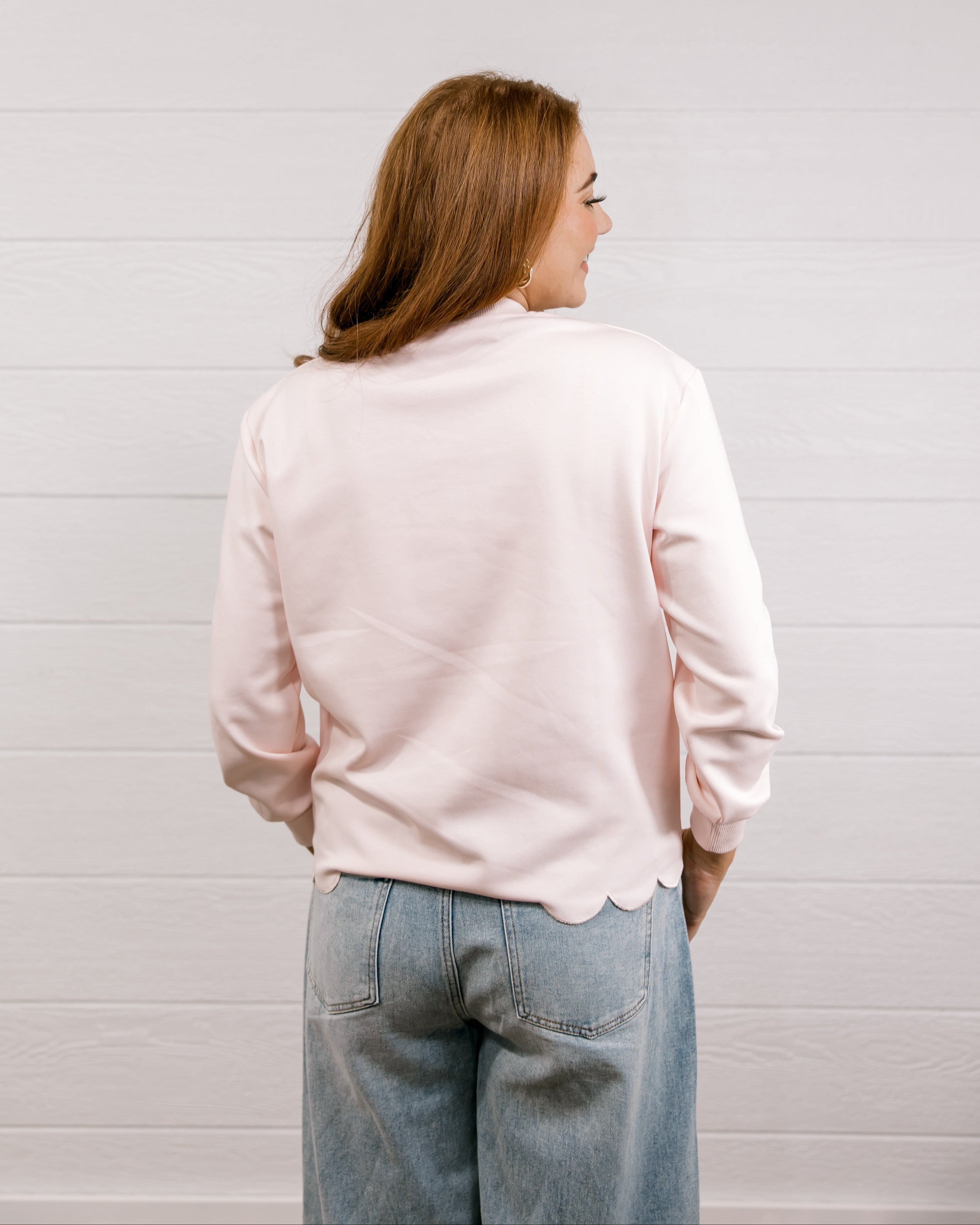 Back view of woman wearing a pink sweatshirt with 'XOXO' embroidered design with scalloped hemline against a white background.