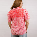 Back view of woman wearing a red patterned blouse that is short sleeve & features red embroidery on front and back against a white background.