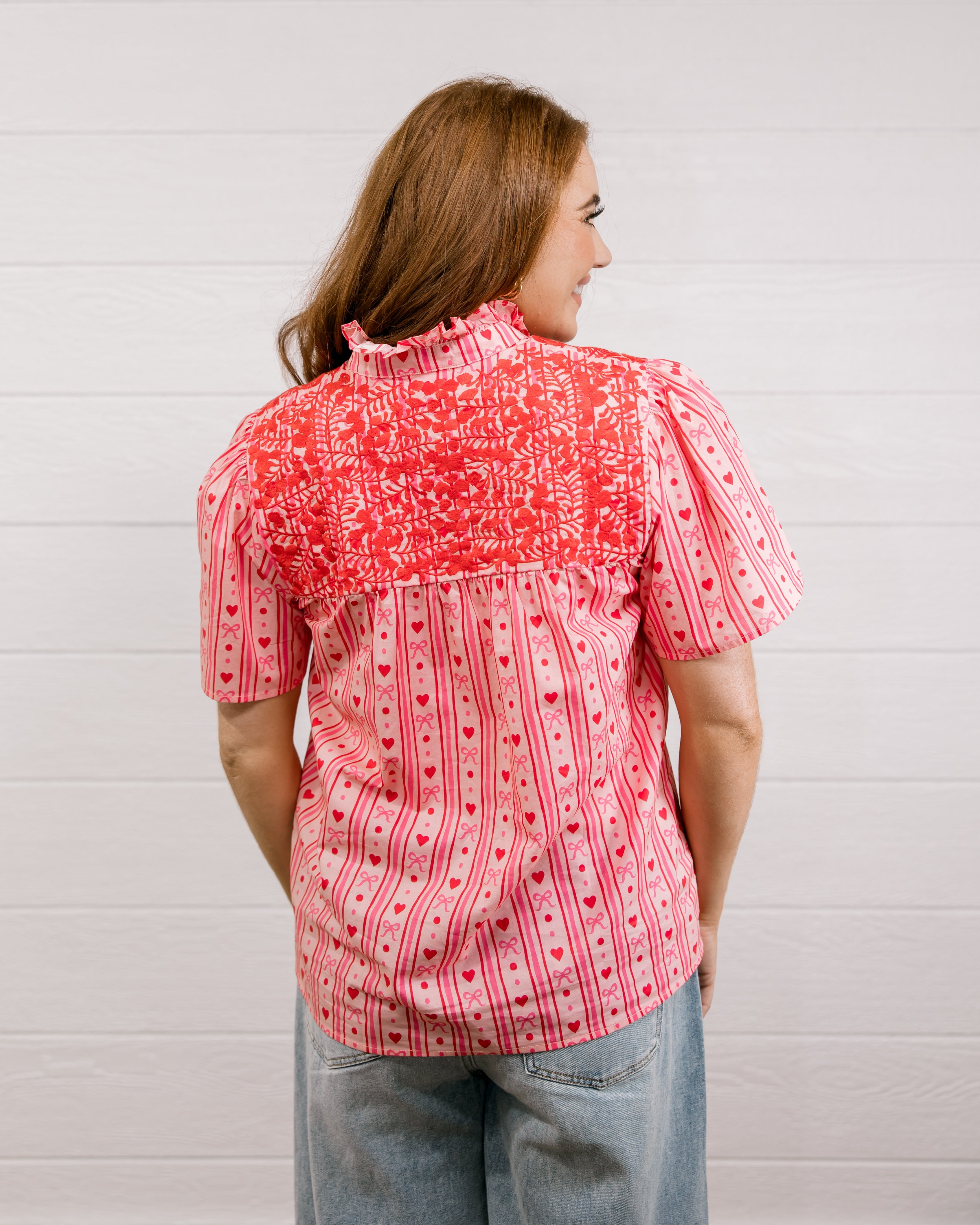 Back view of woman wearing a red patterned blouse that is short sleeve & features red embroidery on front and back against a white background.