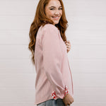 Side view of woman wearing a red blouse that features red & white embroidery on collar & cuff with rolled-up sleeves against a white wall