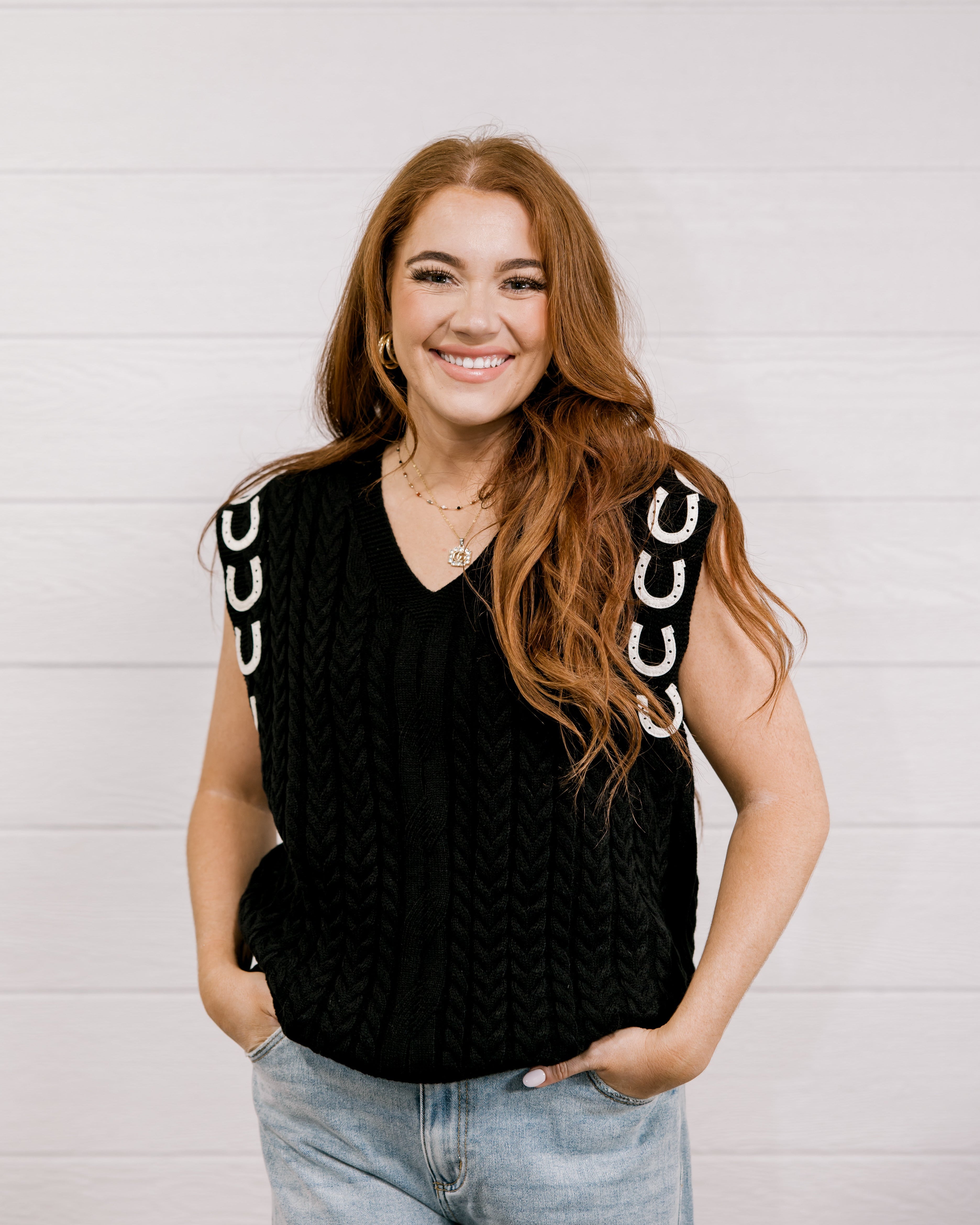 Women wearing a black sweater vest with horse shoes against a white wall.