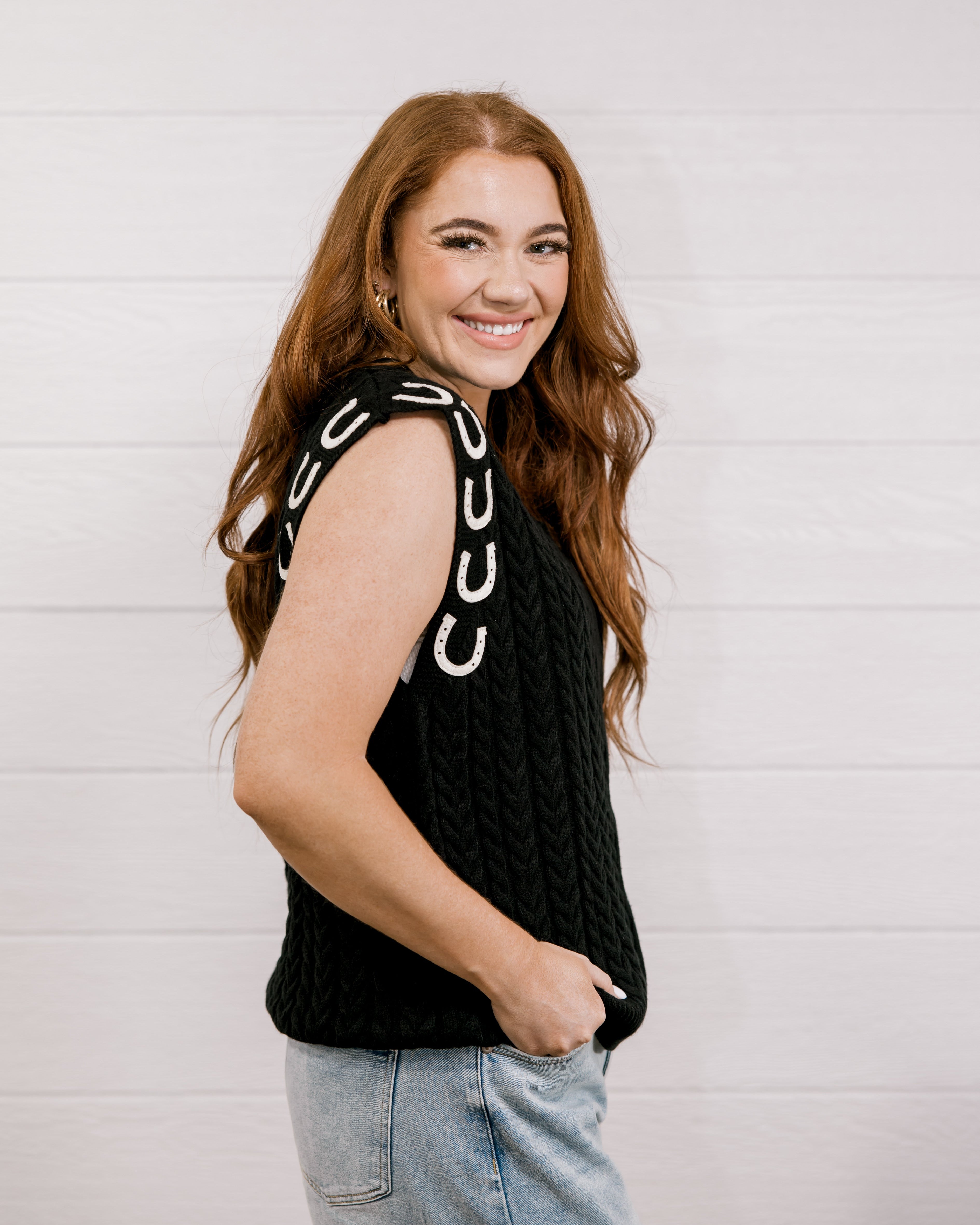 Side view Women wearing a black sweater vest with horse shoes against a white wall.