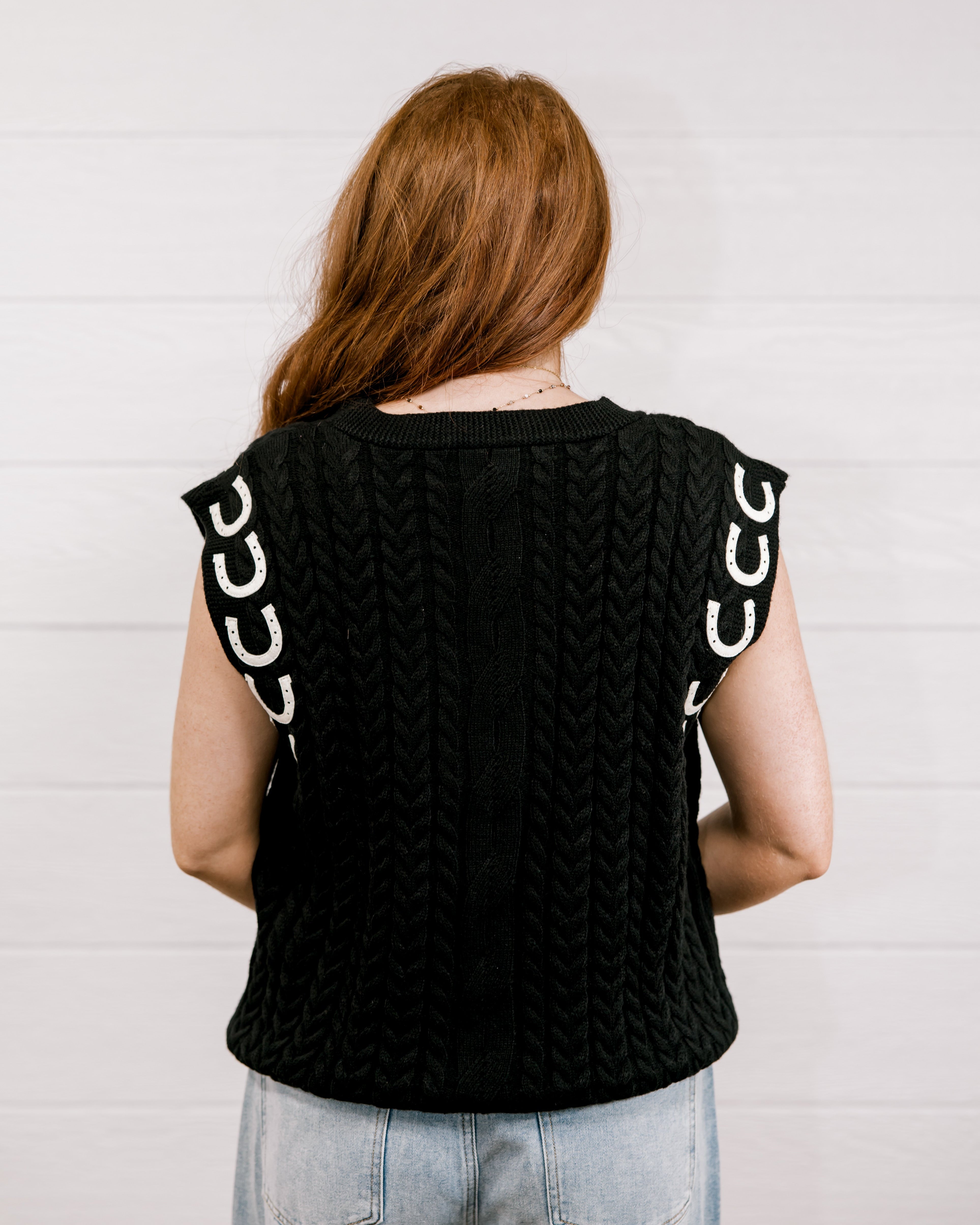 Back view Women wearing a black sweater vest with horse shoes against a white wall.