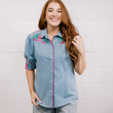 Women wearing a denim pearl snap top with pink western floral embroidery and green leaves. 