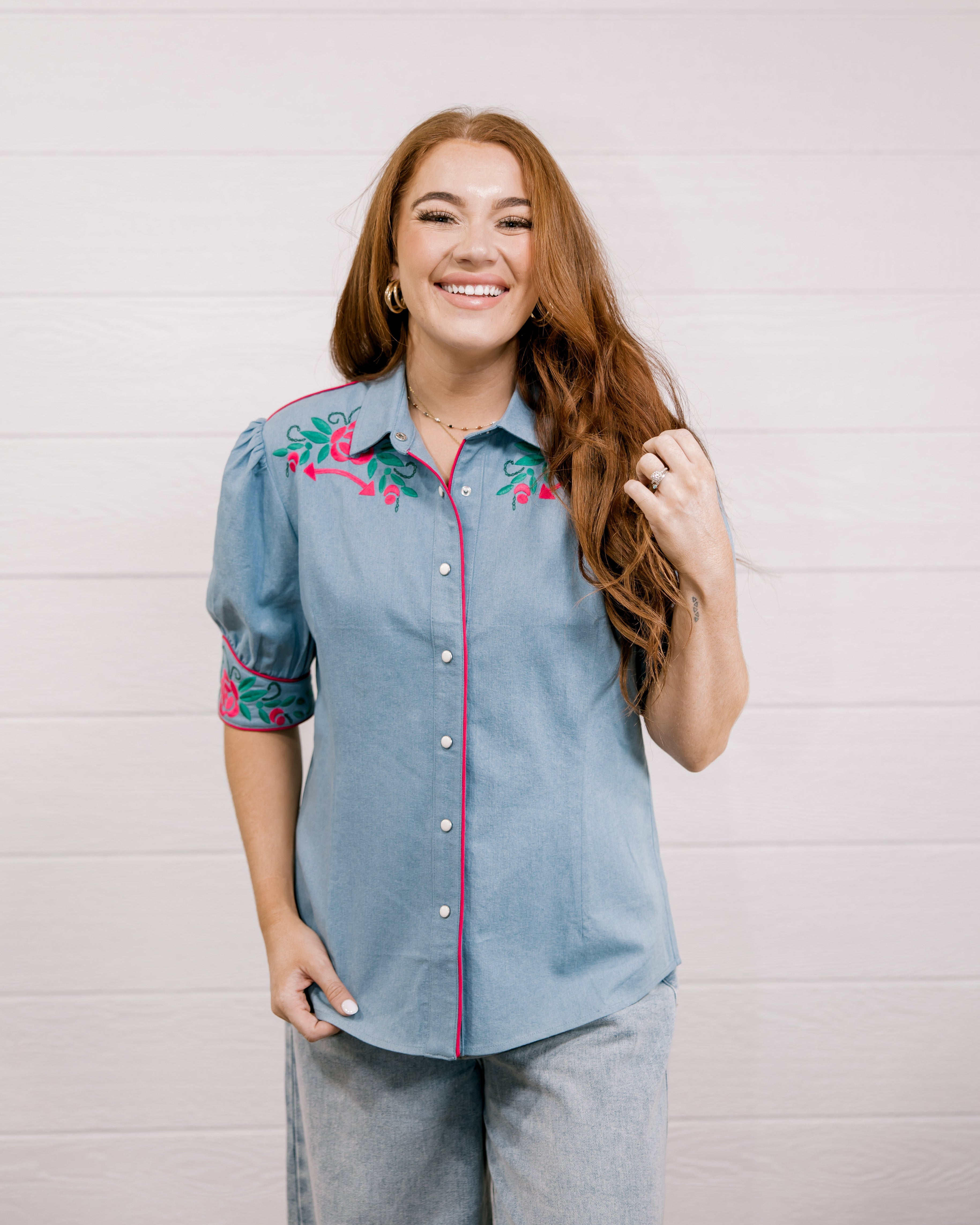 Women wearing a denim pearl snap top with pink western floral embroidery and green leaves. 