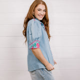 Side view of women wearing a denim pearl snap top with pink western floral embroidery and green leaves. 