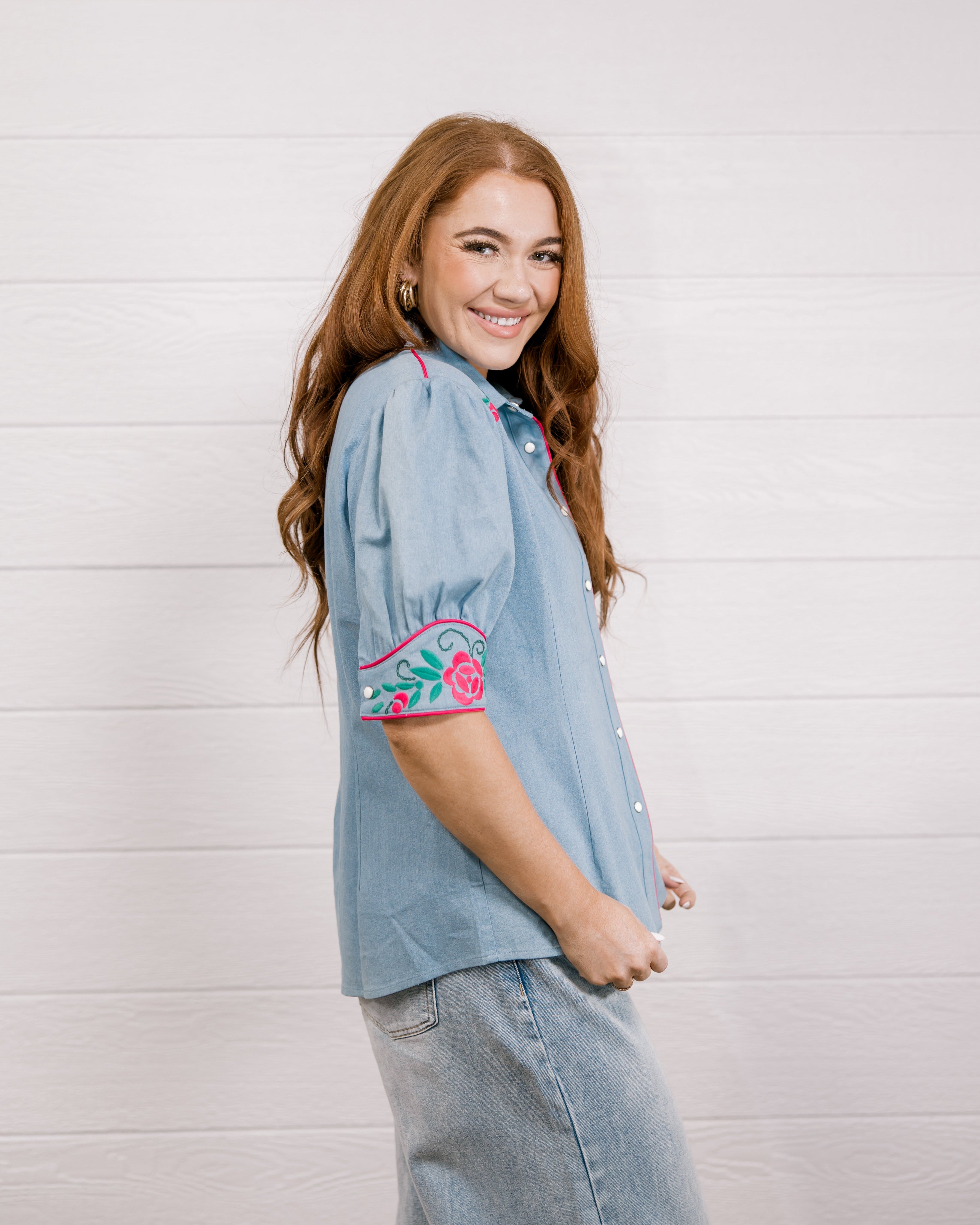 Side view of women wearing a denim pearl snap top with pink western floral embroidery and green leaves. 