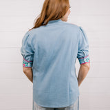 Back view women wearing a denim pearl snap top with pink western floral embroidery and green leaves. 