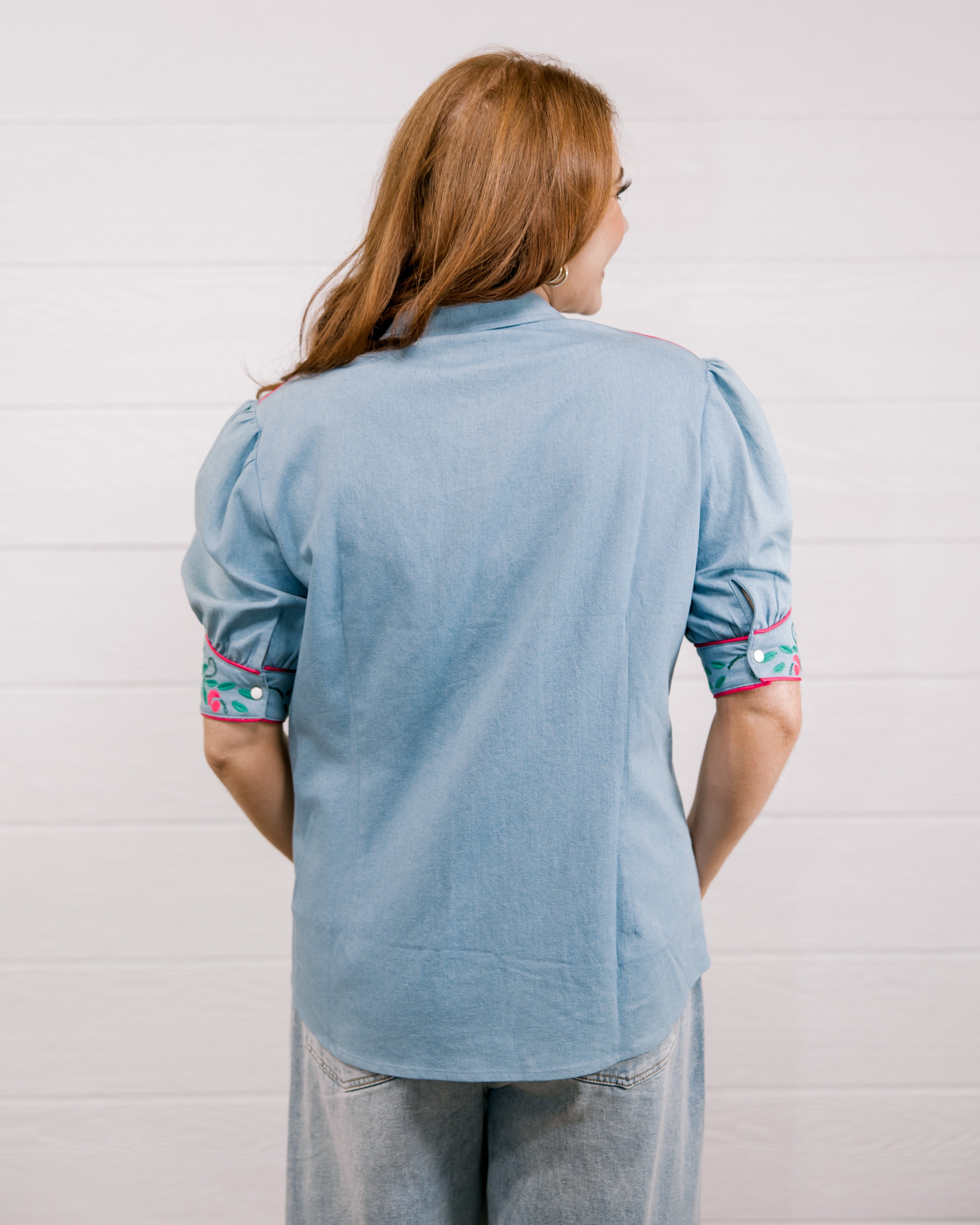 Back view women wearing a denim pearl snap top with pink western floral embroidery and green leaves. 