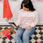 Woman sitting on a checkered floor reading a book, wearing a pink sweatshirt with 'XOXO' text.