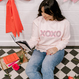 Woman sitting on a checkered floor reading a book, wearing a pink sweatshirt with 'XOXO' text.
