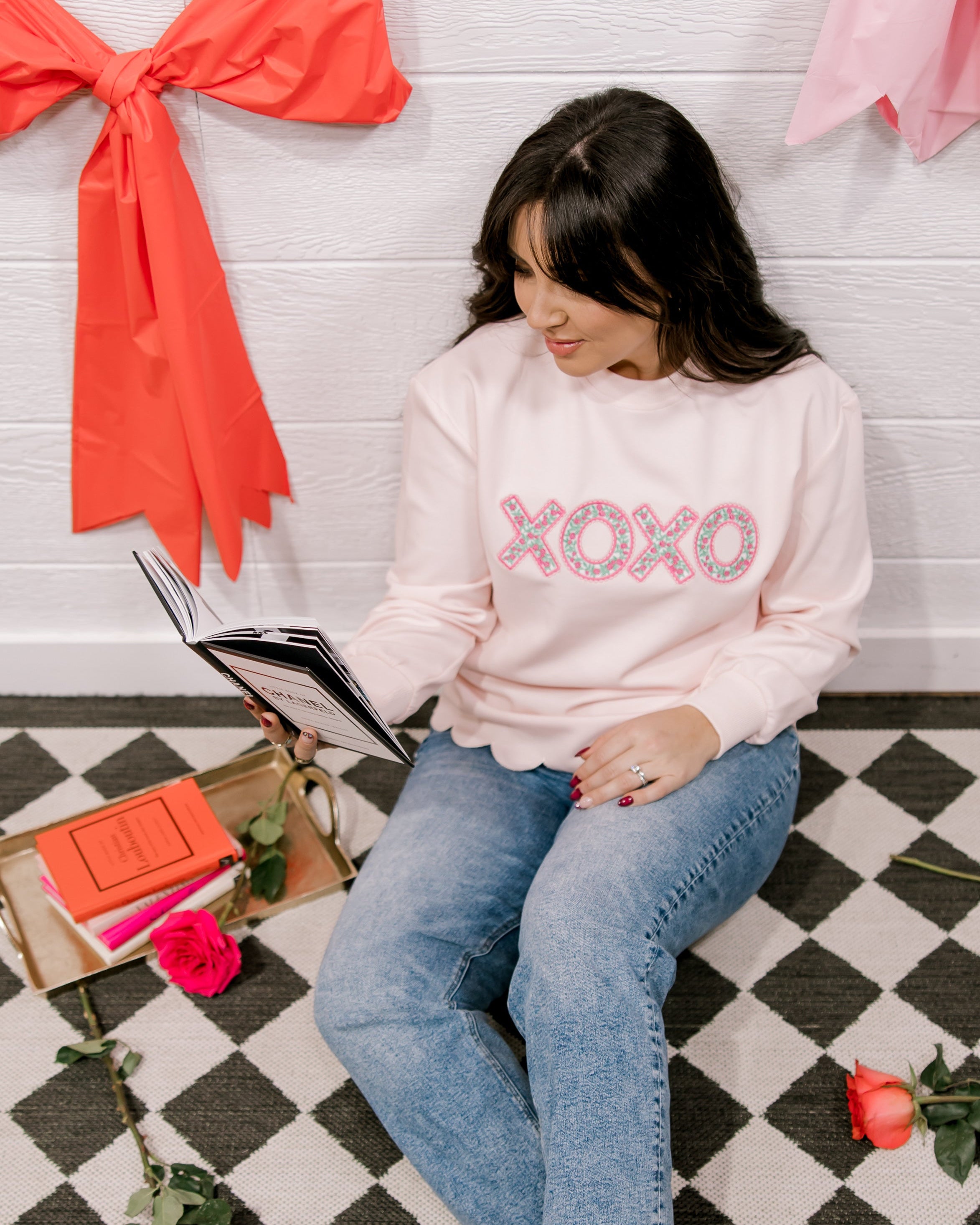 Woman sitting on a checkered floor reading a book, wearing a pink sweatshirt with 'XOXO' text.