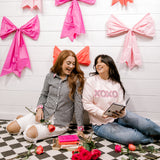 Two women sitting on a checkered floor with decorative bows on the wall behind them.