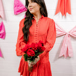 Woman in a red dress holding flowers against a white wall with pink bows.
