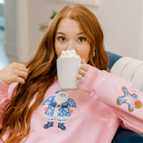 Girl sipping hot coco sitting on a couch in a pink sweatshirt with a blue & white Santa appliqué on the front. 