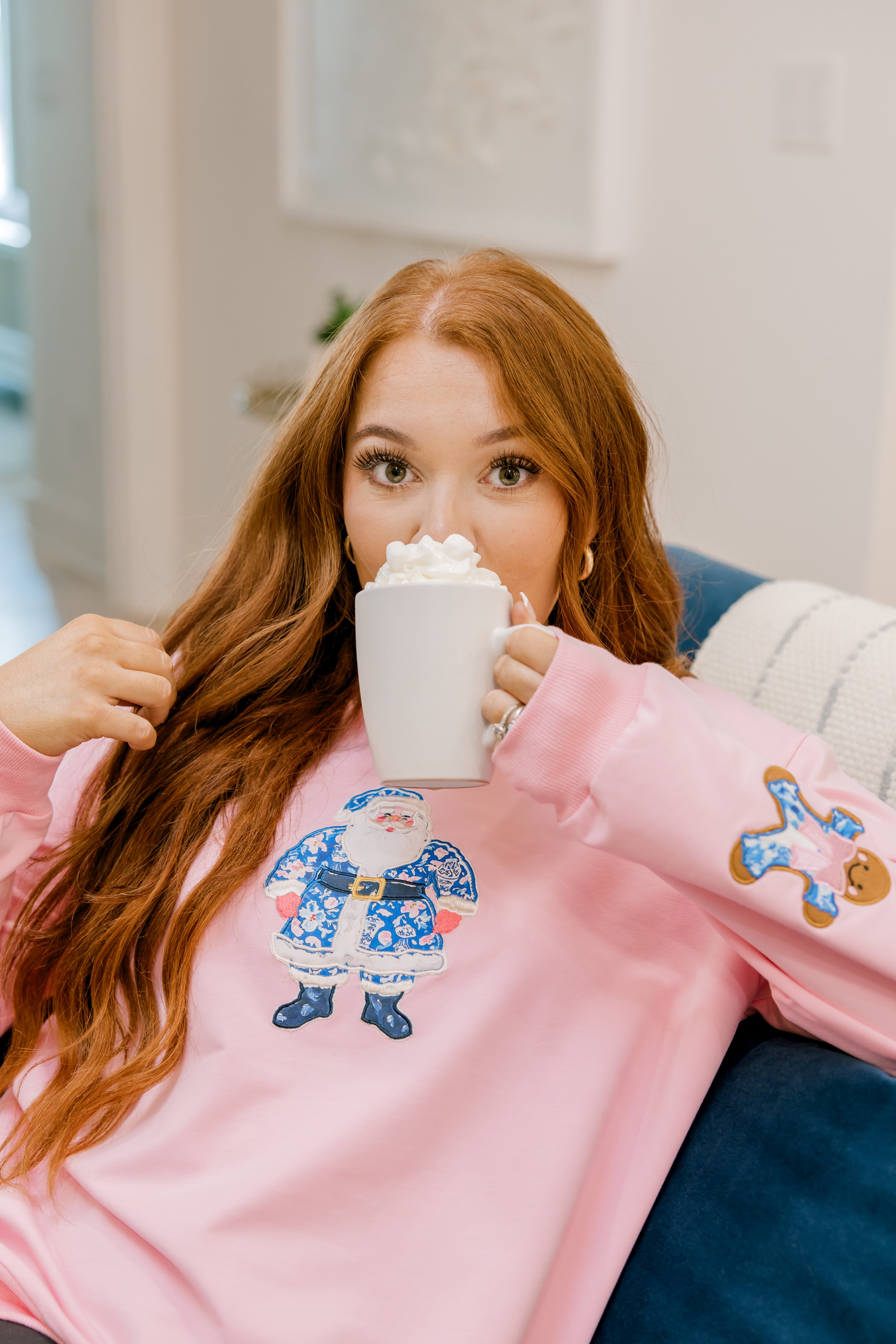 Girl sipping hot coco sitting on a couch in a pink sweatshirt with a blue & white Santa appliqué on the front. 