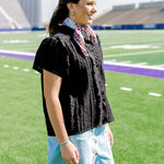 Woman with dark hair standing to the side wearing a short-sleeve black eyelet patterned top, and a light-wash denim skirt.