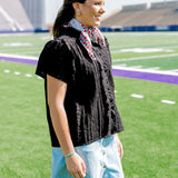 Woman with dark hair standing to the side wearing a short-sleeve black eyelet patterned top, and a light-wash denim skirt.