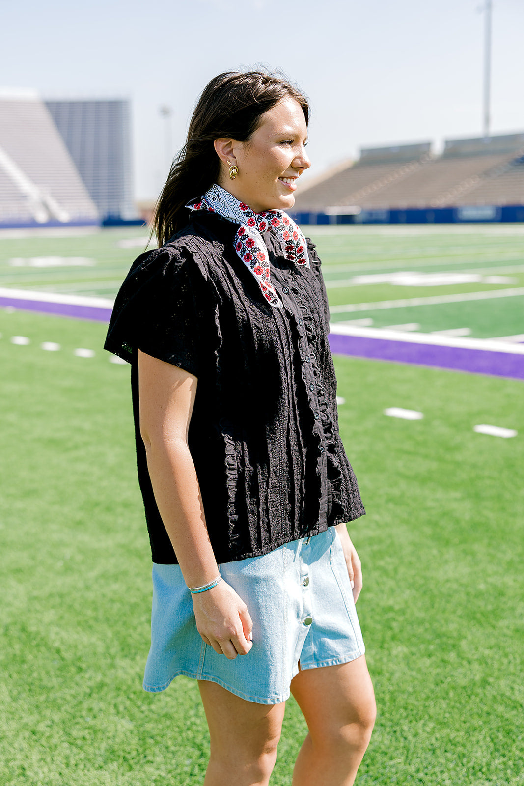 Woman with dark hair standing to the side wearing a short-sleeve black eyelet patterned top, and a light-wash denim skirt.