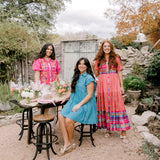 Women in bright colored outfits in a garden having a tea party. 