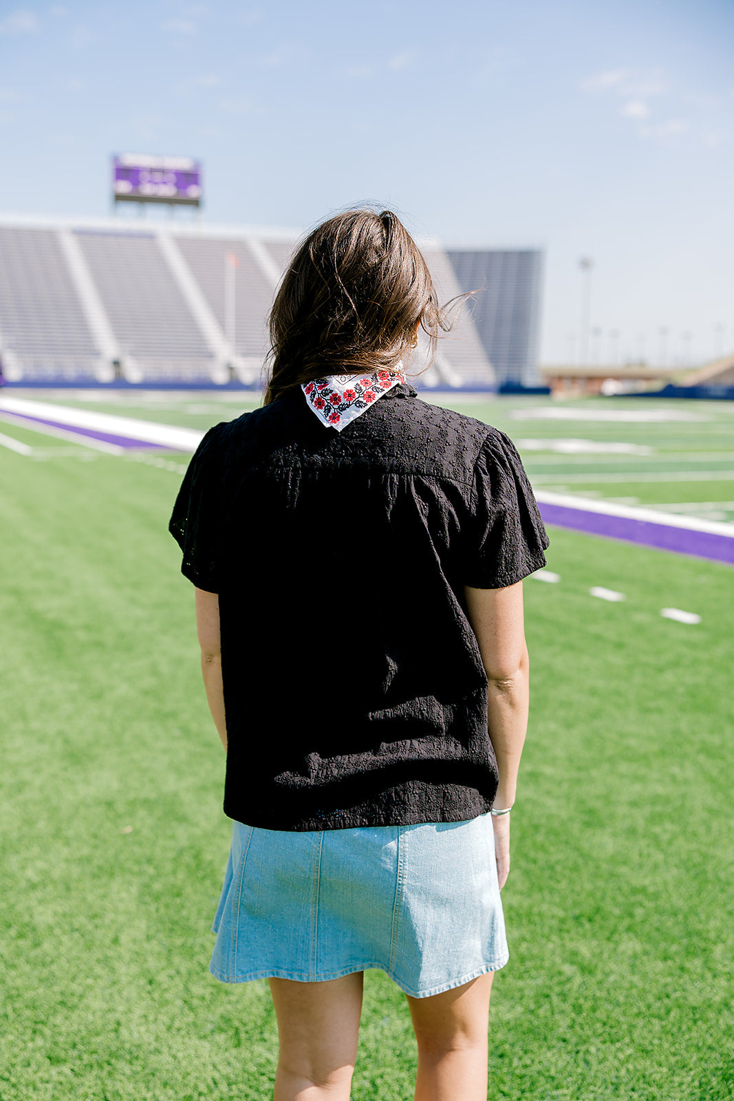 Woman with dark hair wearing a short-sleeve black eyelet pattern top, and a light-wash denim skirt. 