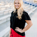 Woman with blonde hair standing in stands wearing a short-sleeve black eyelet patterned top, and a red denim skirt. 