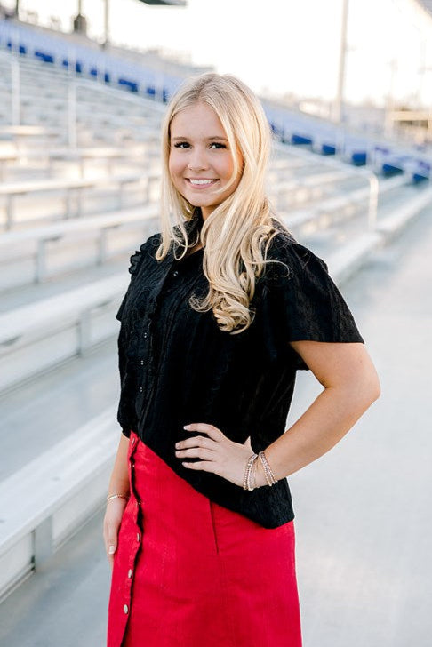Woman with blonde hair standing in stands wearing a short-sleeve black eyelet patterned top, and a red denim skirt. 