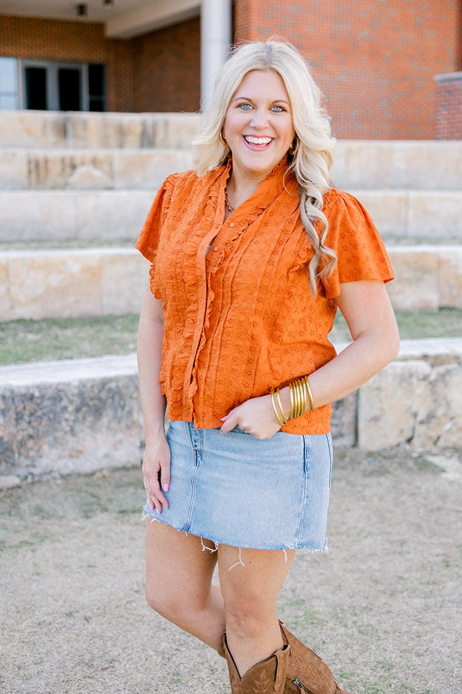 Woman with blonde hair standing in a front profile position wearing a short-sleeve burnt orange eyelet top and a light-wash denim skirt.