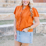 Woman with blonde hair standing in a front profile position wearing a short-sleeve burnt orange eyelet top and a light-wash denim skirt.