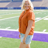 Woman with blonde hair standing in a side profile position wearing a short-sleeve eyelet top in burnt orange. The model paired this top with light-wash denim shorts. 