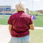 Woman with blonde hair standing in a back profile position wearing the Lilly Top in a lifestyle scene. The focal point of this image is the maroon eyelet pattern as well as pleating on back and ruffle on collar. The model paired this top with light-wash denim shorts. 