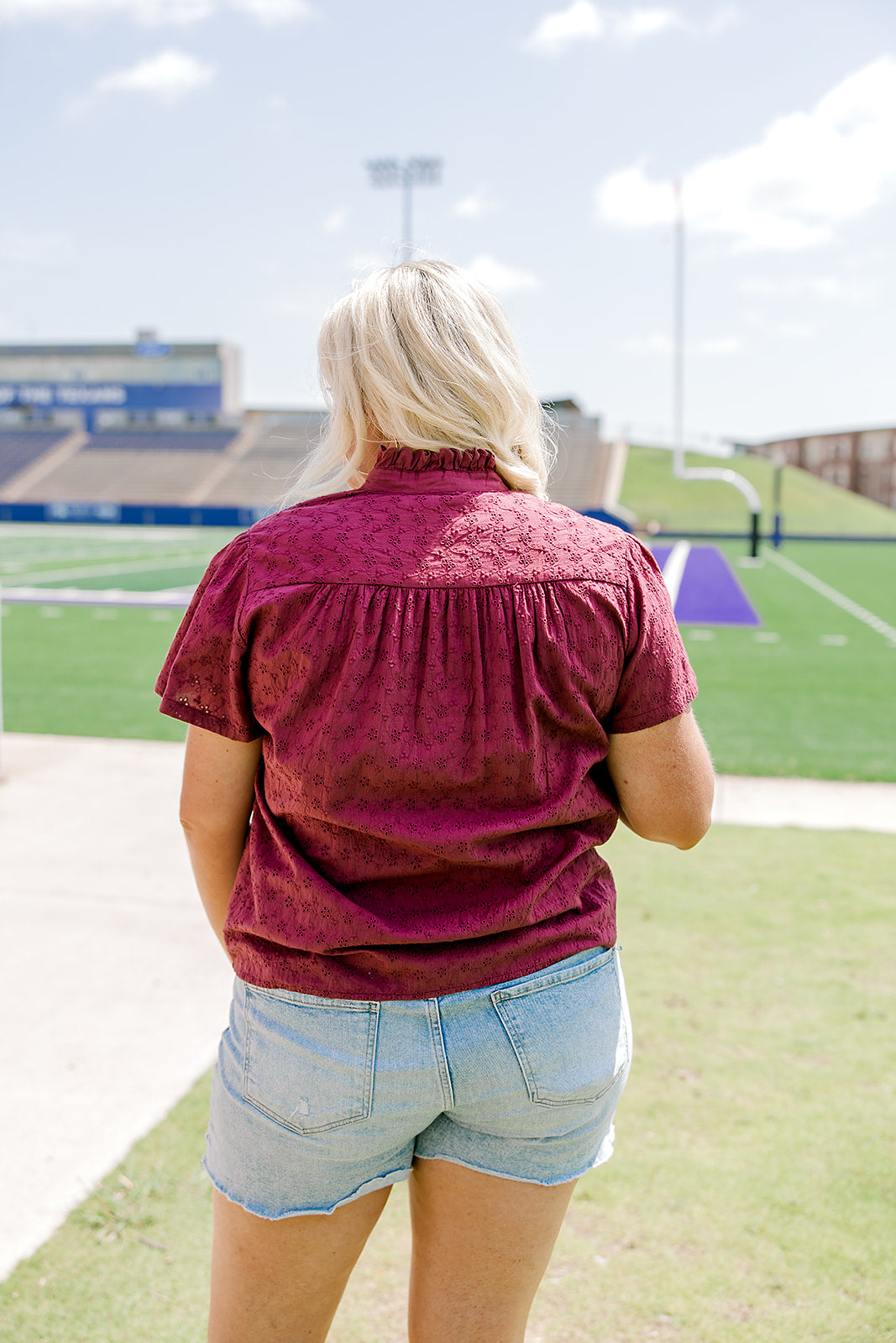Woman with blonde hair standing in a back profile position wearing the Lilly Top in a lifestyle scene. The focal point of this image is the maroon eyelet pattern as well as pleating on back and ruffle on collar. The model paired this top with light-wash denim shorts. 