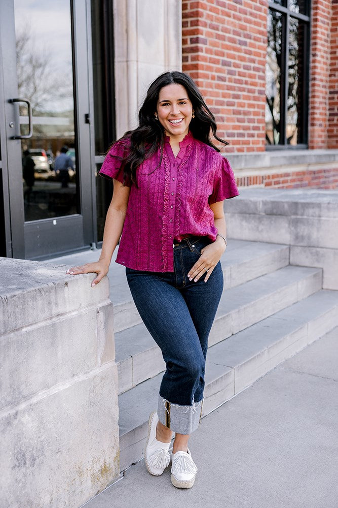 Woman with dark hair standing in a front profile position wearing the Lilly Top on the steps of campus. The focal point of this image is the ruffle down side of buttons as well as eyelet pattern covering top. The model paired this top with dark-wash denim jeans. 