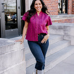 Woman with dark hair standing in a front profile position wearing the Lilly Top on the steps of campus. The focal point of this image is the ruffle down side of buttons as well as eyelet pattern covering top. The model paired this top with dark-wash denim jeans. 