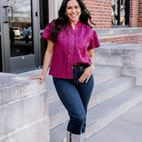 Woman with dark hair standing in a front profile position wearing the Lilly Top on the steps of campus. The focal point of this image is the ruffle down side of buttons as well as eyelet pattern covering top. The model paired this top with dark-wash denim jeans. 
