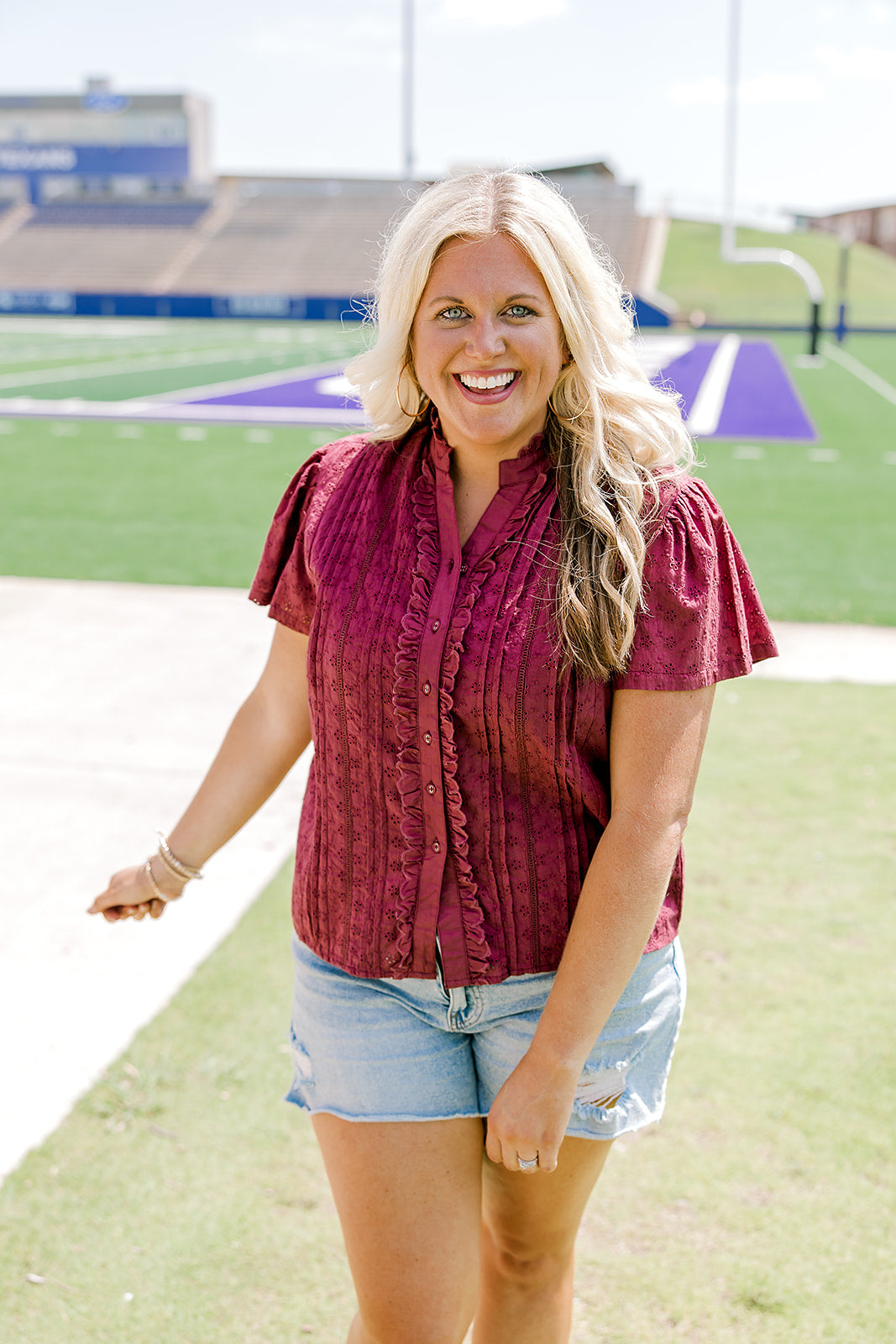 Woman with blonde hair standing in a front profile position wearing the Lilly Top in a lifestyle scene. The focal point of this image is the maroon eyelet pattern across top, ruffles along wooden buttons and high collar with ruffles. The model paired this top with light-wash denim shorts. 