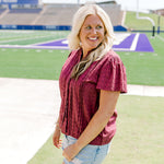 Woman with blonde hair standing in a side profile position wearing the Lilly Top in a lifestyle scene. The focal point of this image is the pleating on sleeve as well as maroon eyelet pattern across top. The model paired this top with light-wash denim shorts. 
