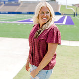 Woman with blonde hair standing in a side profile position wearing the Lilly Top in a lifestyle scene. The focal point of this image is the pleating on sleeve as well as maroon eyelet pattern across top. The model paired this top with light-wash denim shorts. 